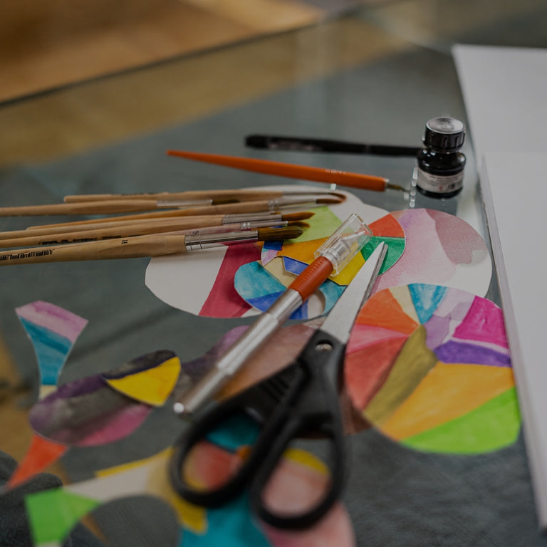A close-up of a glass table holding a colorful children's art project with painted paper circles, several wooden paintbrushes, a small bottle of ink, and black-handled scissors.