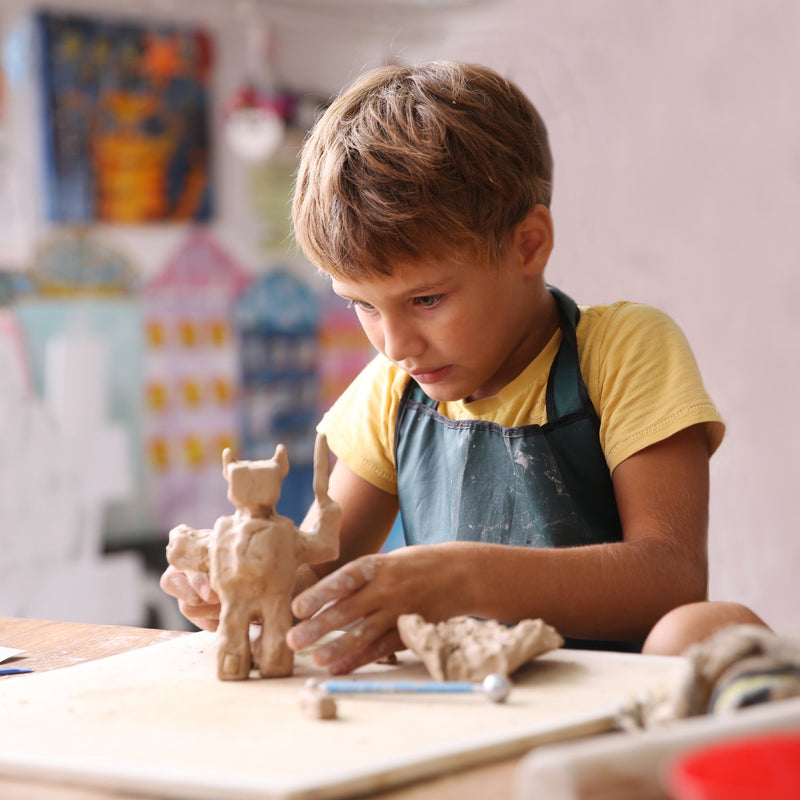 A young boy with brown hair, wearing a yellow t-shirt and a dark blue apron, sculpting a small clay animal on a wooden table.