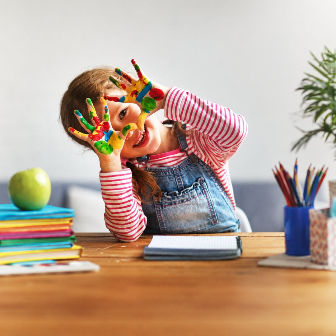 A young child with colorful hand-painted fingers holds them up to their face while sitting at a wooden desk with school supplies, including a green apple, notebooks, and a cup of colored pencils.
