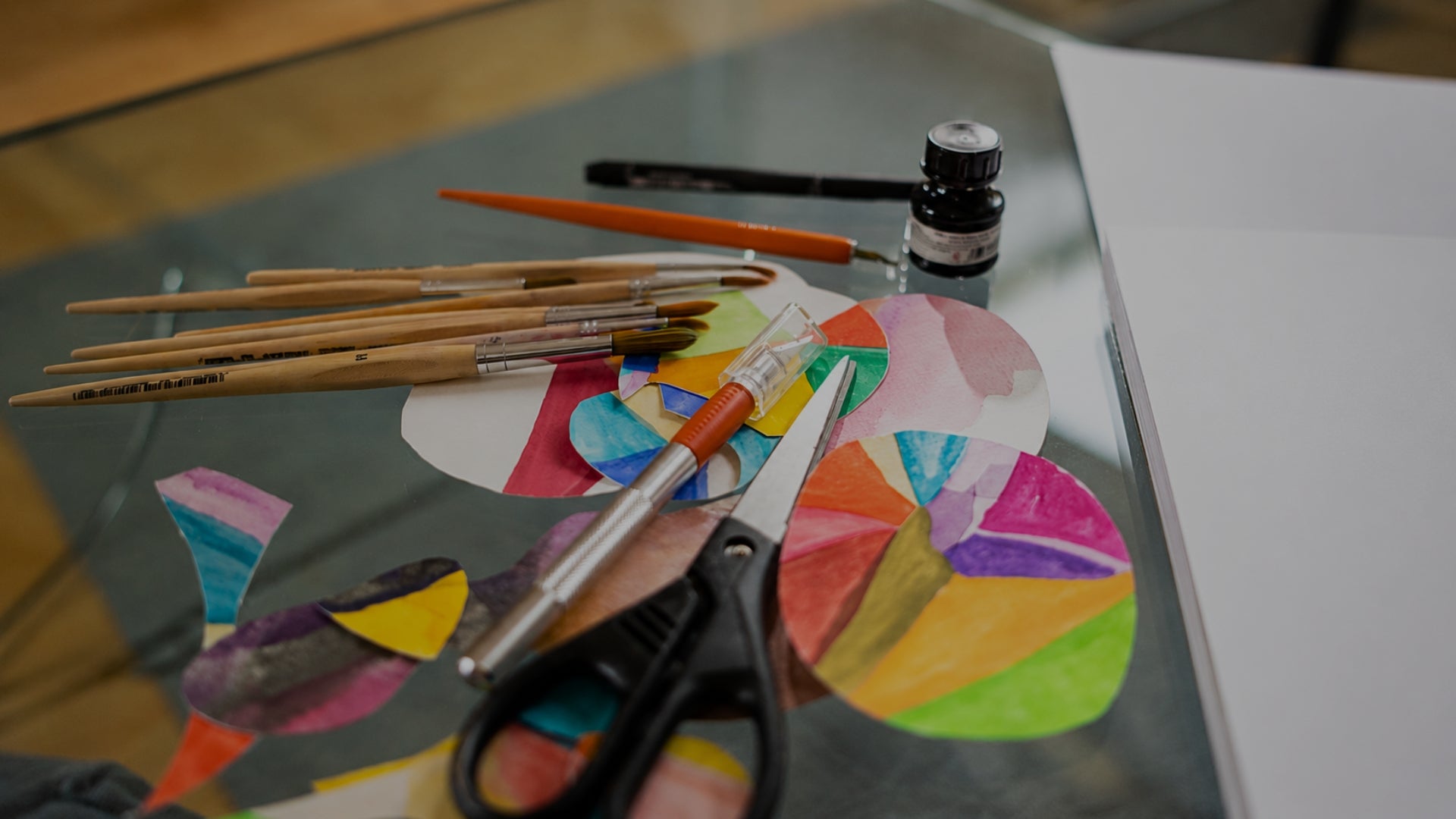 A close-up of a glass table holding a colorful children's art project with painted paper circles, several wooden paintbrushes, a small bottle of ink, and black-handled scissors.