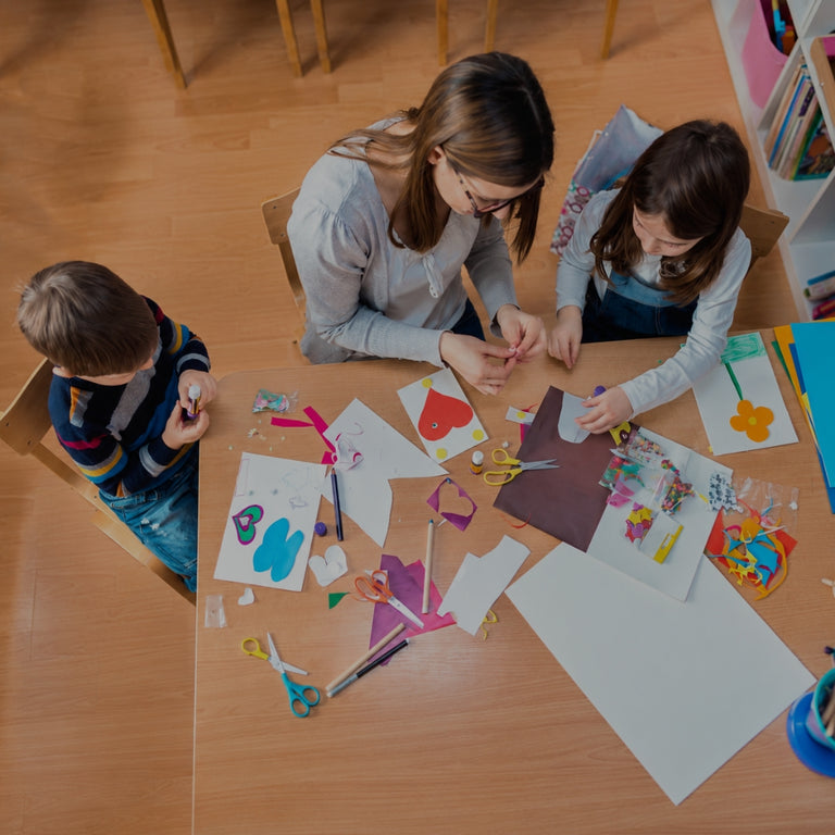 Three children sitting around a wooden table engaged in a craft activity, surrounded by colorful paper cutouts, scissors, and art supplies in a brightly lit indoor room.