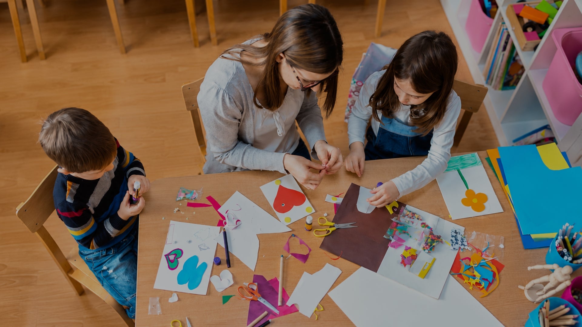 Three children sitting around a wooden table engaged in a craft activity, surrounded by colorful paper cutouts, scissors, and art supplies in a brightly lit indoor room.