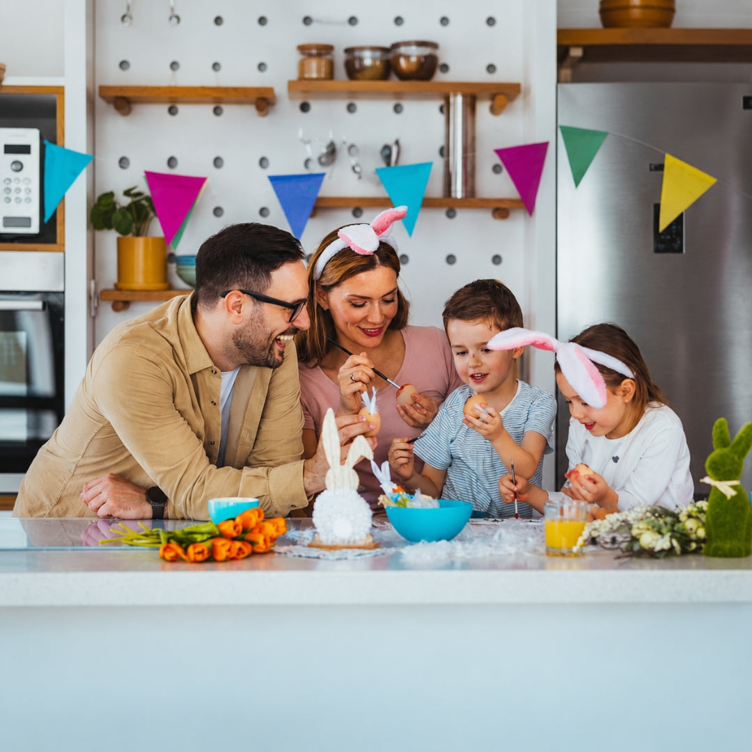 A family of four celebrates Easter in a bright kitchen, wearing pink bunny ears headbands and gathered around a table with an egg-crafting project, carrots, and bowls.