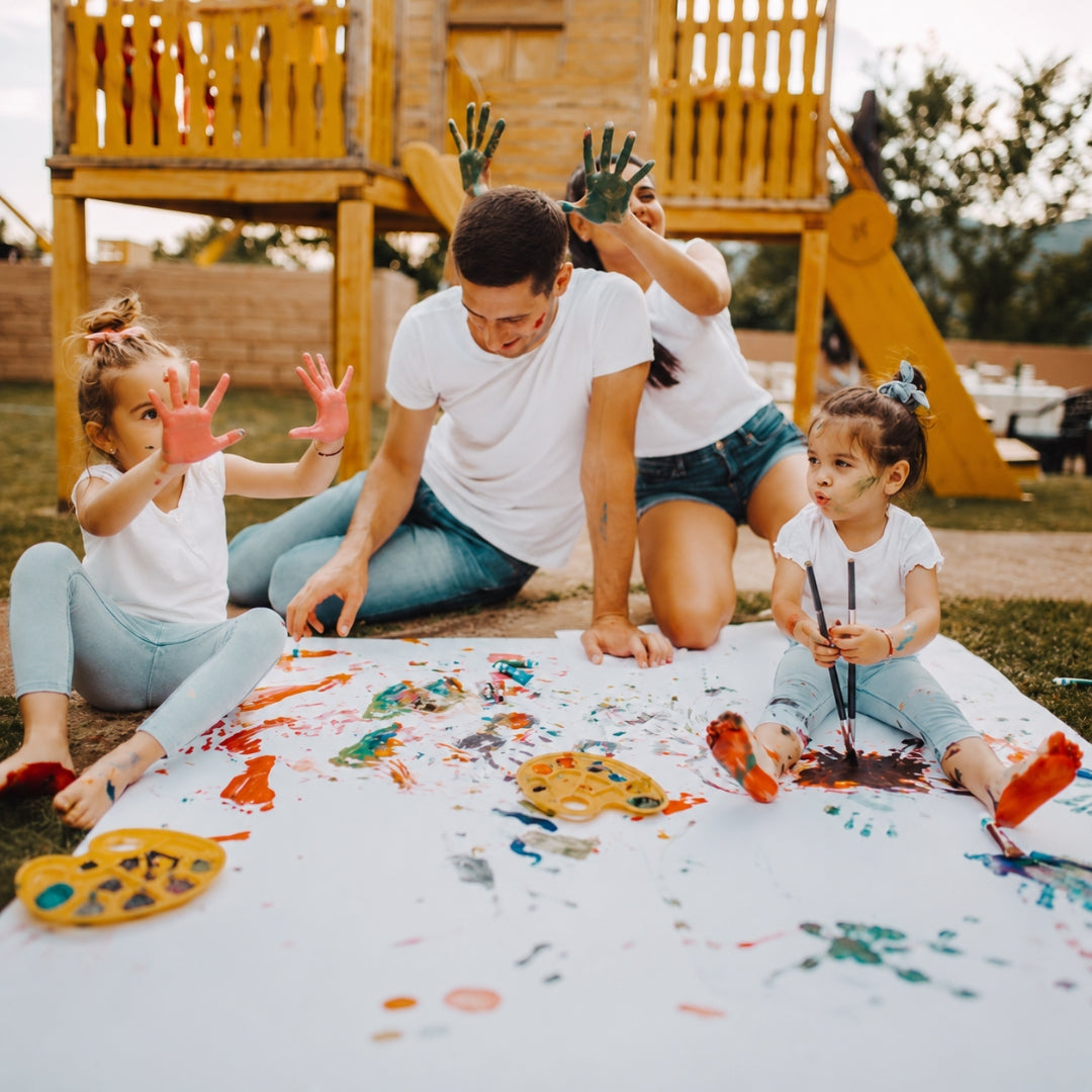 A family of four enjoys outdoor painting on a white sheet on the grass in front of a wooden playground. The father and mother are kneeling down, while the two young girls sit beside them, all covered in colorful paint splatters.