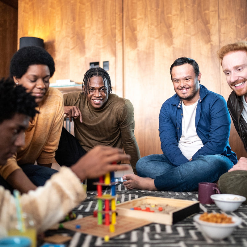 A group of four friends laughing and playing a board game in a cozy, warmly lit living room setting with wooden walls and tables holding snacks and drinks.