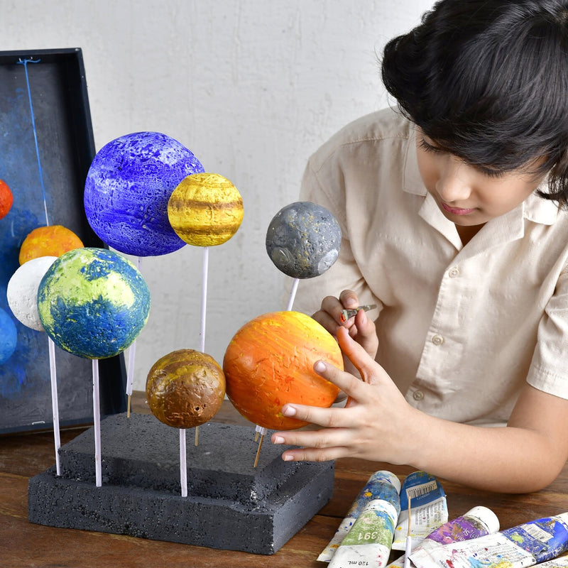A person painting a colorful solar system model set of planets on a black display base. The set includes planets painted in blue, green, yellow, orange, and gray, with paint tubes on a table in the foreground.
