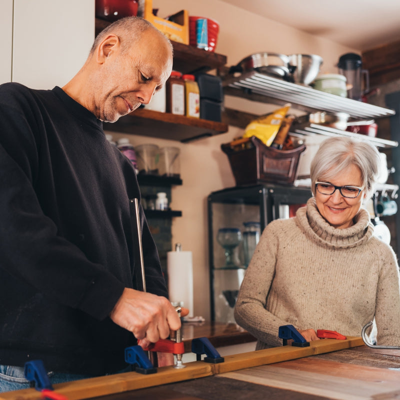 An older man in a black long-sleeve shirt and an older woman in a beige turtleneck sweater are working together on a wooden board in a kitchen setting. The man is using a clamp with a wooden handle, and both are focused on the task at hand.
