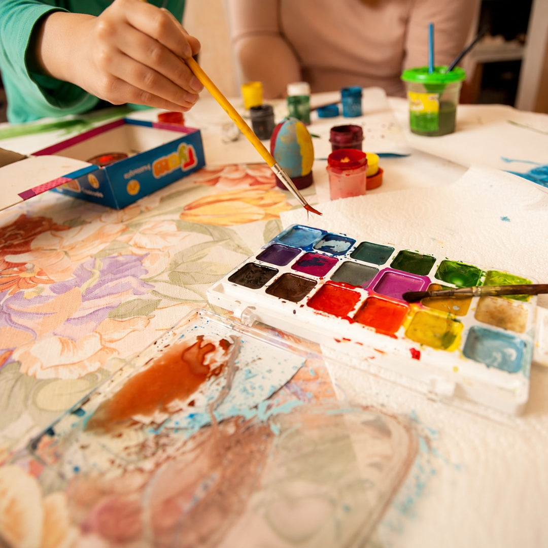 A close-up of a person's hand holding a paintbrush over a watercolor palette filled with multiple colors, on a table covered with painting supplies and a floral watercolor painting in progress.