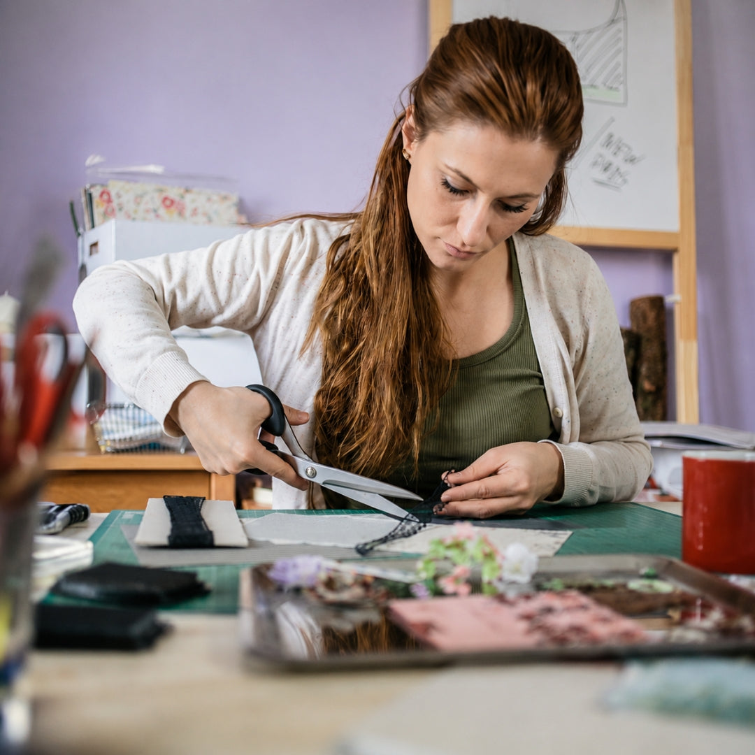 A woman with long light brown hair is seated at a table, using black-handled scissors to cut a black ribbon or cord. She wears a beige cardigan over an olive green top.
