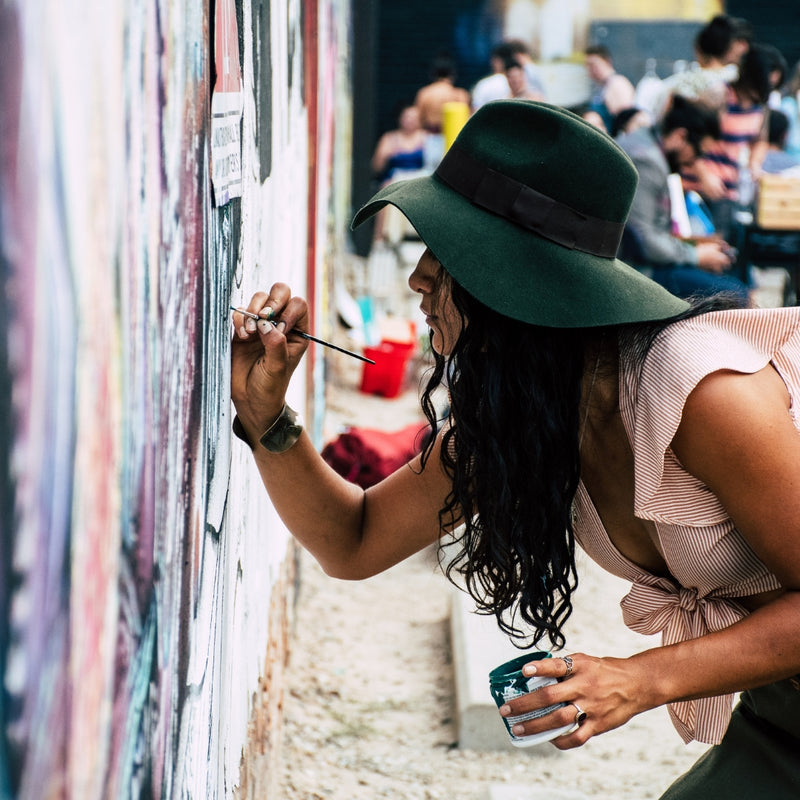 A woman wearing a wide-brimmed dark green hat and a light pink ruffled top leans against a wall, painting a mural with a brush while holding a cup in an outdoor street setting.