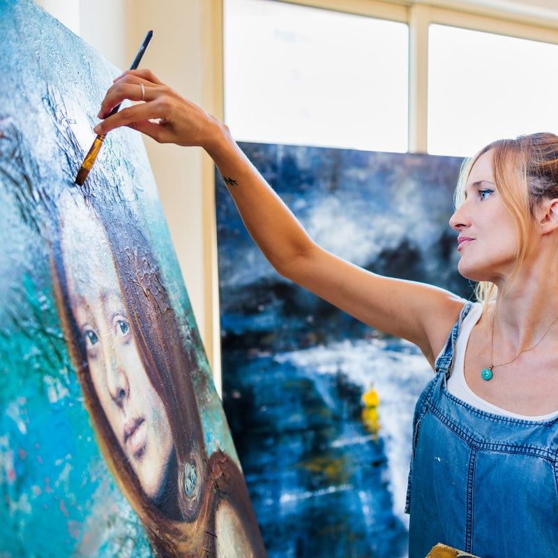 A woman painting a portrait on a canvas in an indoor studio setting, with a large blue-toned artwork leaning behind her.