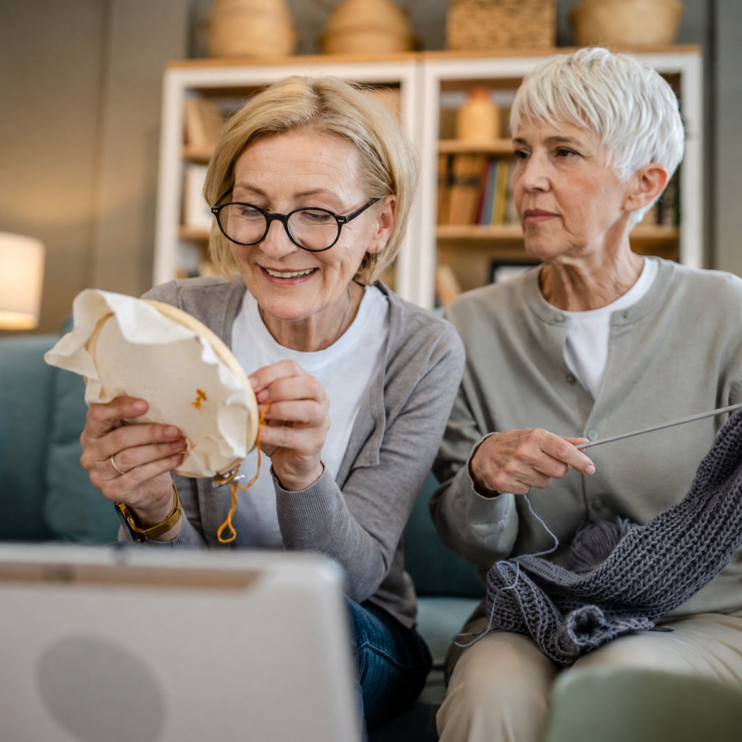 Two women sitting on a couch in a cozy living room setting, one holding a knitted fabric sample and looking at a laptop, while the other holds a knitting needle and yarn.
